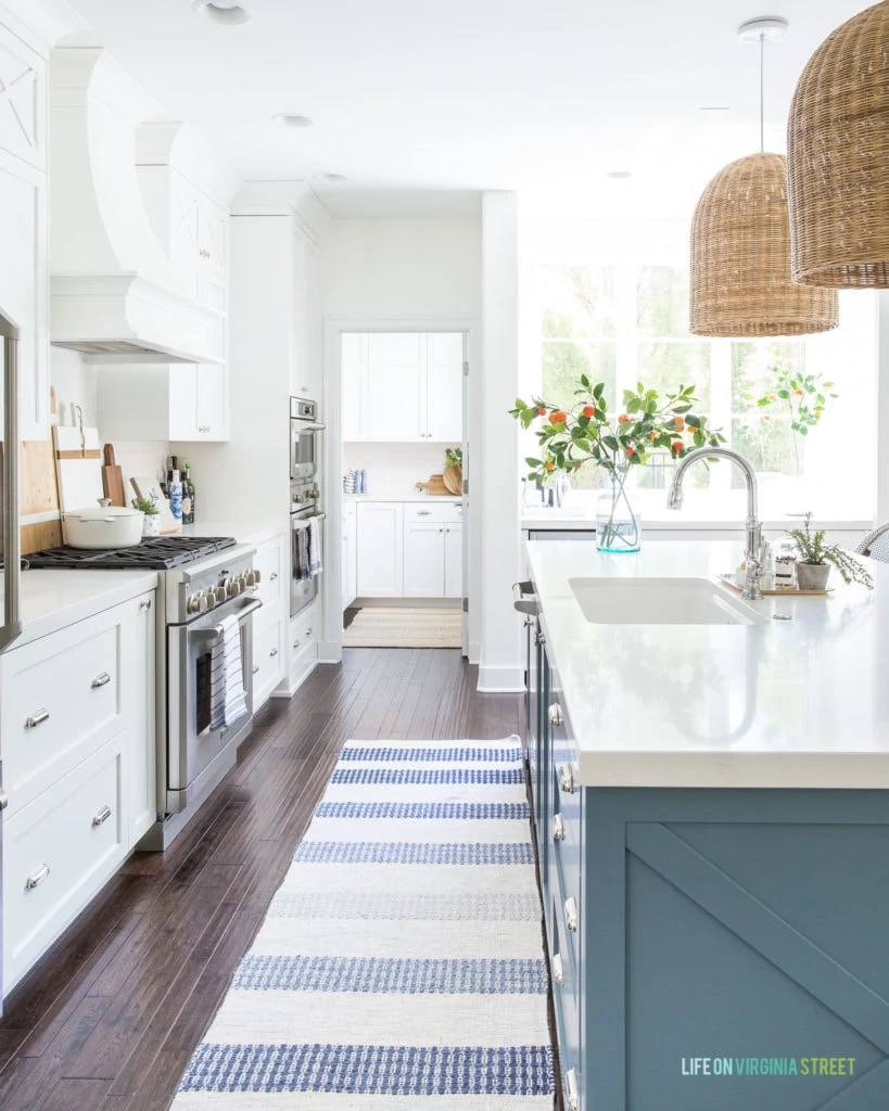 A kitchen with a blue island and white cabinets. There is a blue and white striped rug, basket pendant light fixtures over the island, and faux orange stems in a vase.