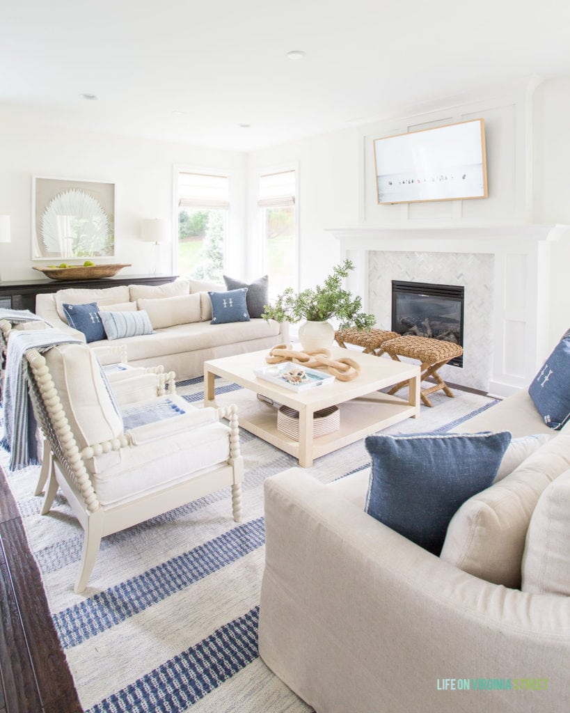 Our modern coastal living room with white spindle armchairs, linen sofas, a blue striped rug, raffia coffee table and fan palm art. The walls are painted Benjamin Moore Simply White.