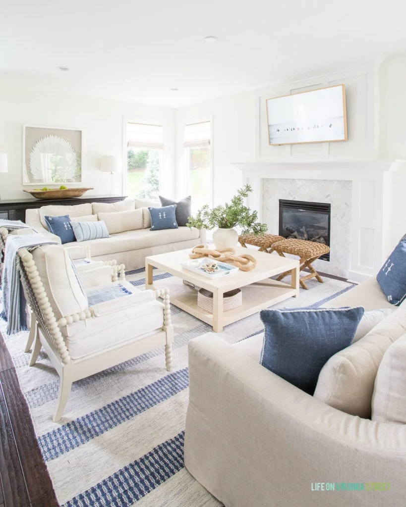 A coastal living room with linen sofas, white spindle chairs, a raffia coffee table, palm art, and blue pillows.