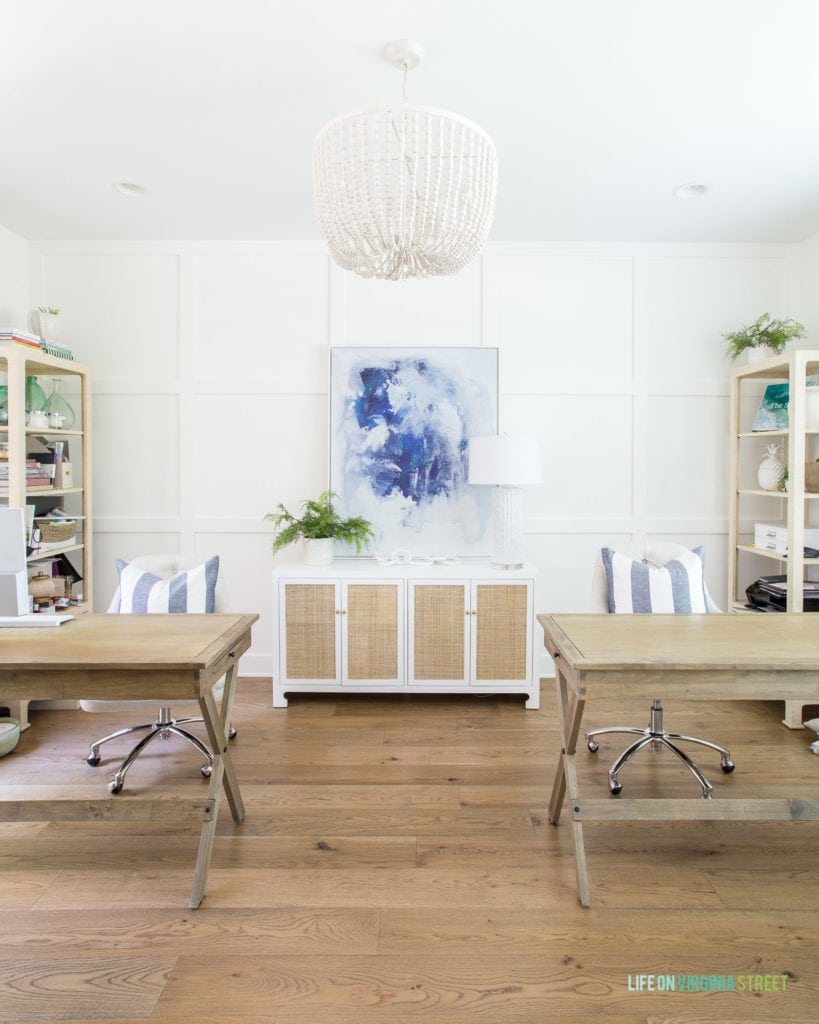 A home office with two wood desks, cane console table, blue abstract art, white bead chandelier and white oak hardwood floors.