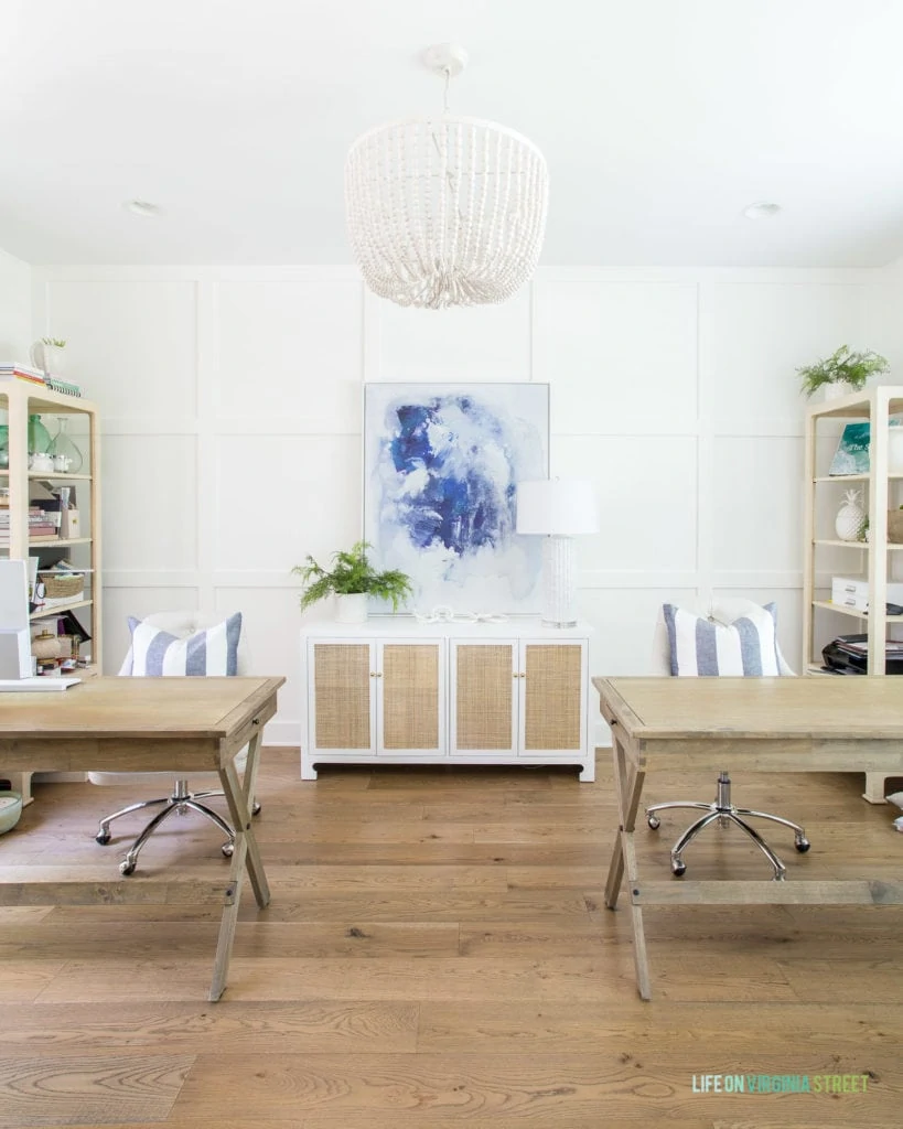 A home office with a white board and batten grid wall, wood desks, white oak hardwood floors, white wood bead chandelier, a cane cabinet, and blue abstract art.