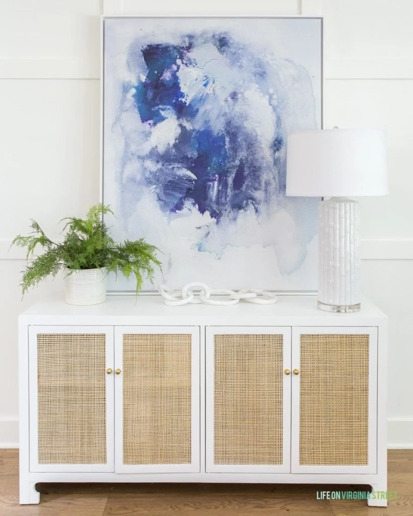 A cane cabinet with blue abstract art, a white lamp, faux fern, and marble chain.