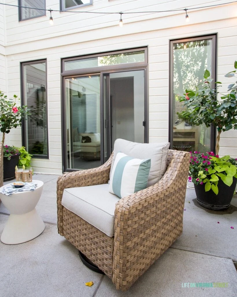 A painted white house with bronze window frames an outdoor courtyard area. Includes woven outdoor chairs, string lights, lantern wall sconces, white concrete side tables and planters.