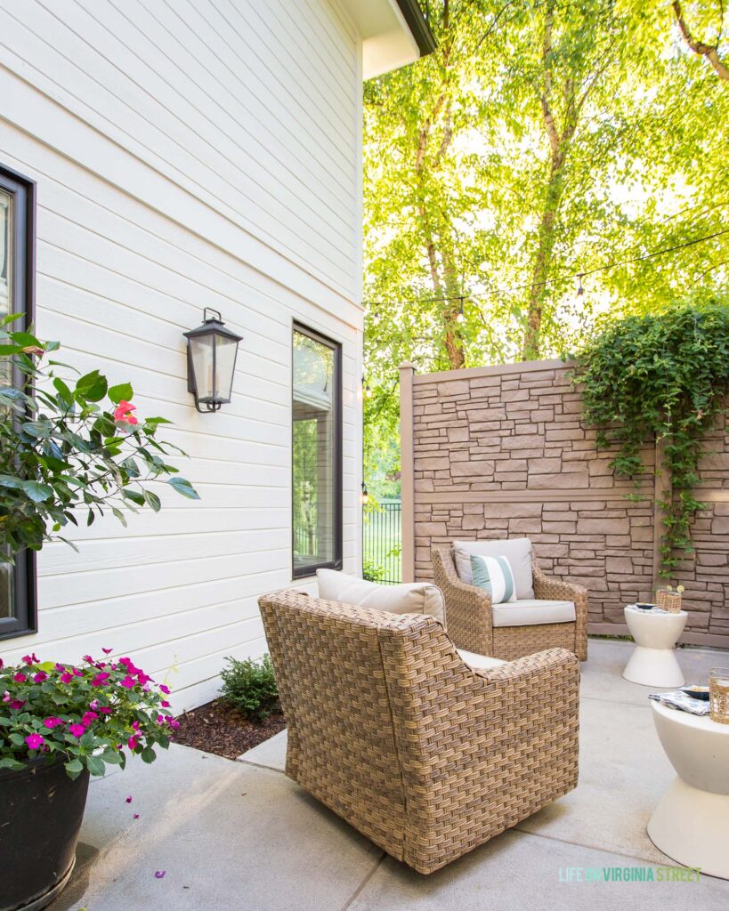An outdoor courtyard area with faux stone walls, wicker chairs, concrete side tables, white house exterior and bronze window frames.