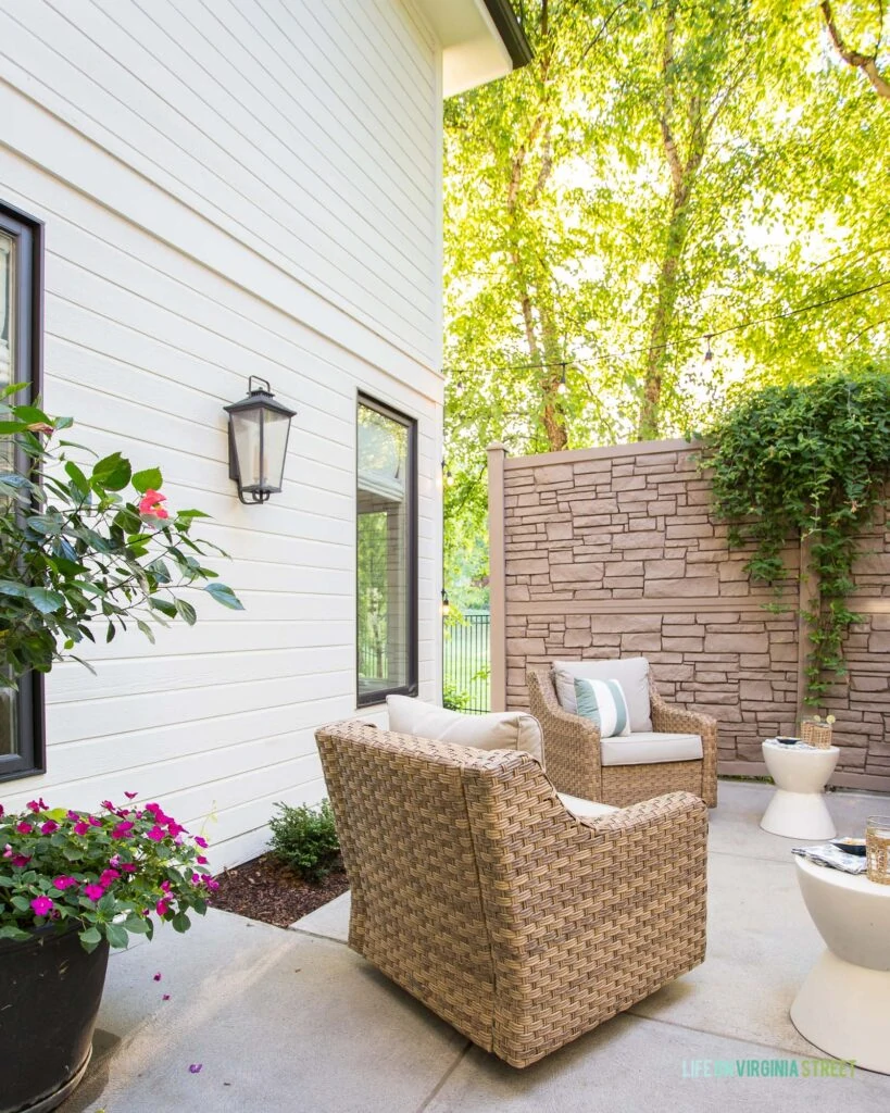 A summer courtyard with faux wicker armchairs, concrete side tables, black outdoor wall lanterns, striped Sunbrella outdoor pillows, and colorful flowers and trees.