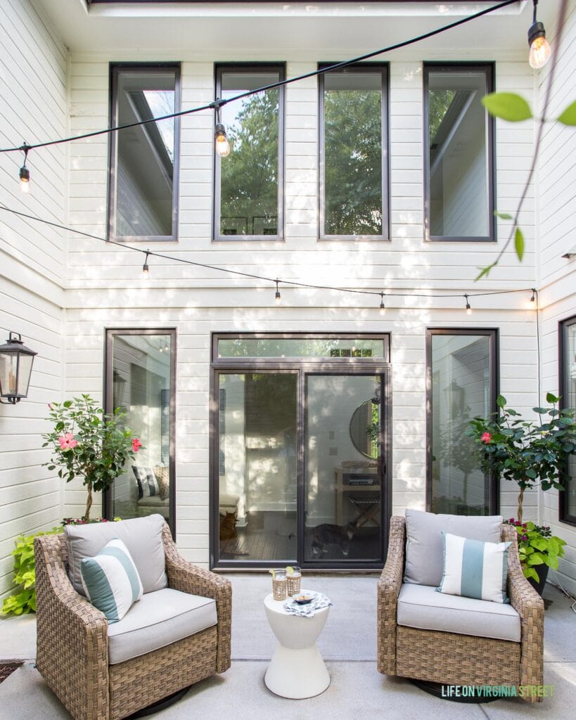 An outdoor courtyard on a white house with black window trim. Includes natural woven swivel chairs, string lights, and outdoor wall lanterns.