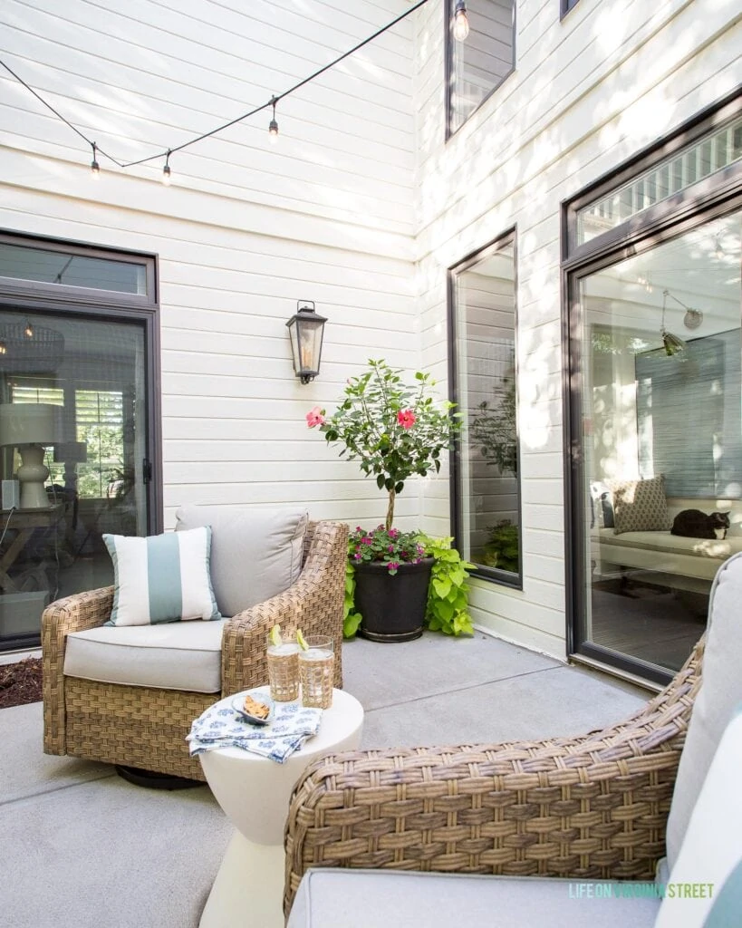 Outdoor wall lights in a courtyard with natural woven chairs, planters with colorful flowers and string lights on the white house.