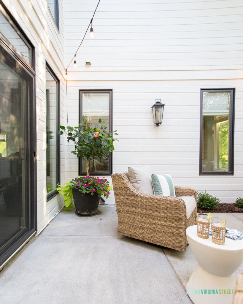 A painted white house with an outdoor courtyard area. Includes woven outdoor chairs, string lights, lantern wall sconces, white concrete side tables and planters.