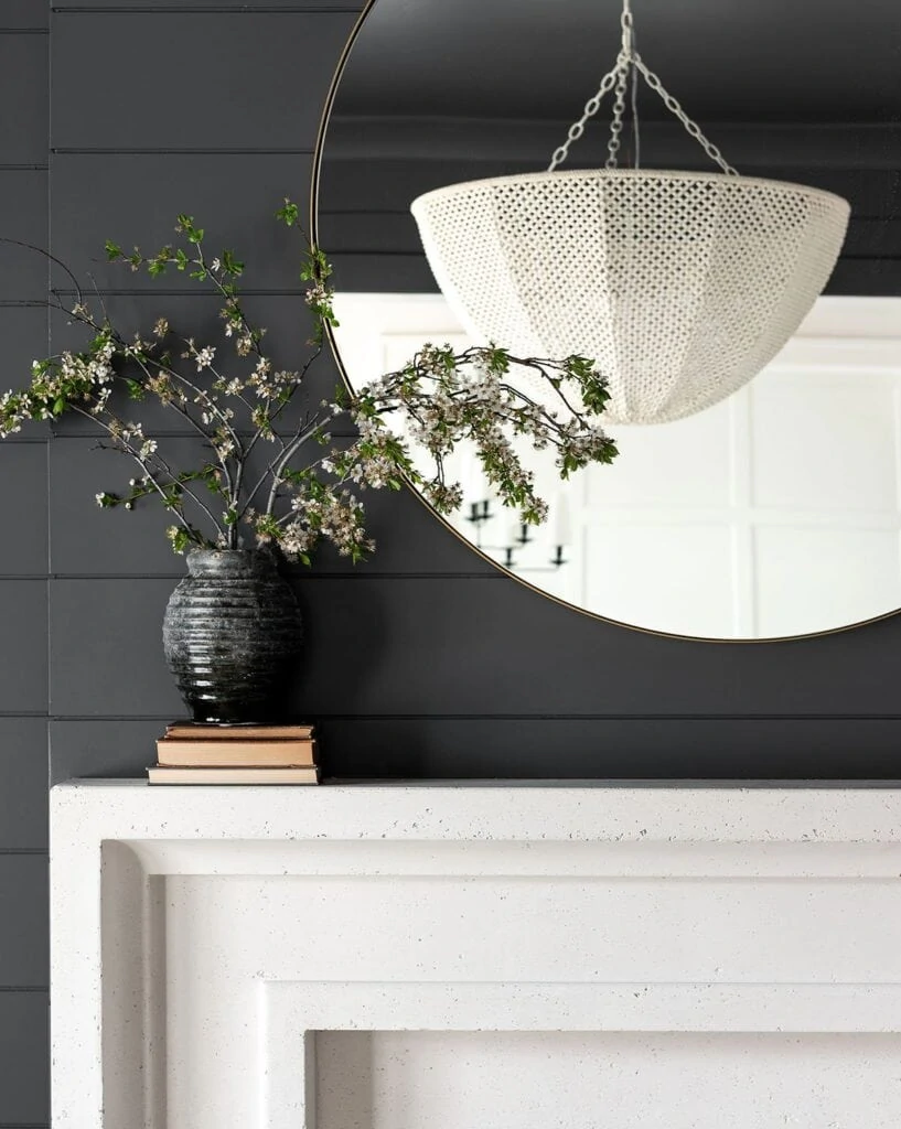 A weathered charcoal vase filled with fresh branches, styled on a limestone fireplace with black shiplap, a round mirror and white chandelier.