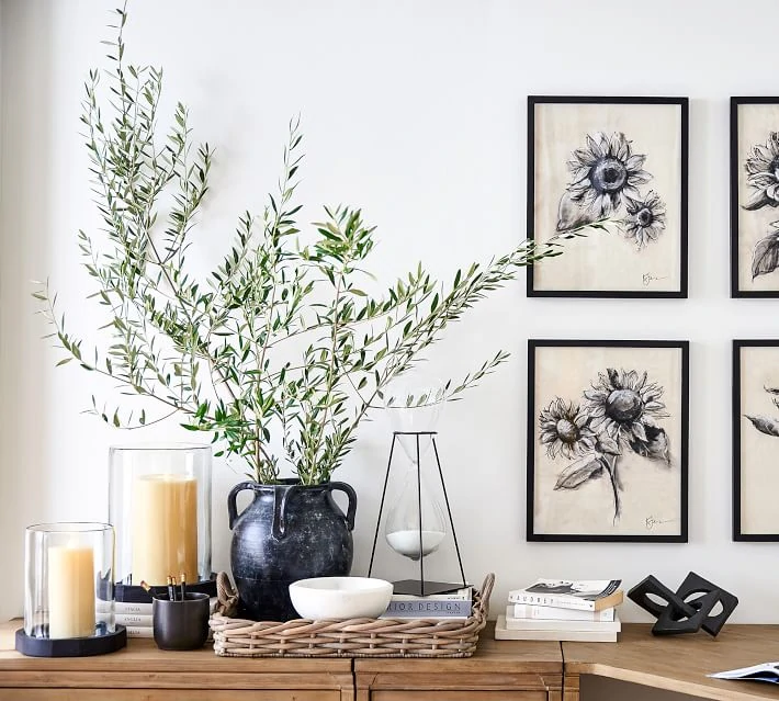 A beautiful weathered black vase filled with olive branches. Also shown are pillar candles and sunflower art styled on a desk.