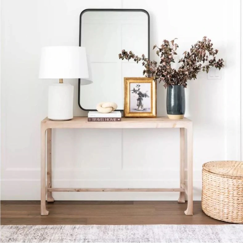 A natural wood console table styled with a white ceramic lamp, tall metal mirror, limestone knot decor, gold framed art, and a dark charcoal vase filled with fall leaves.