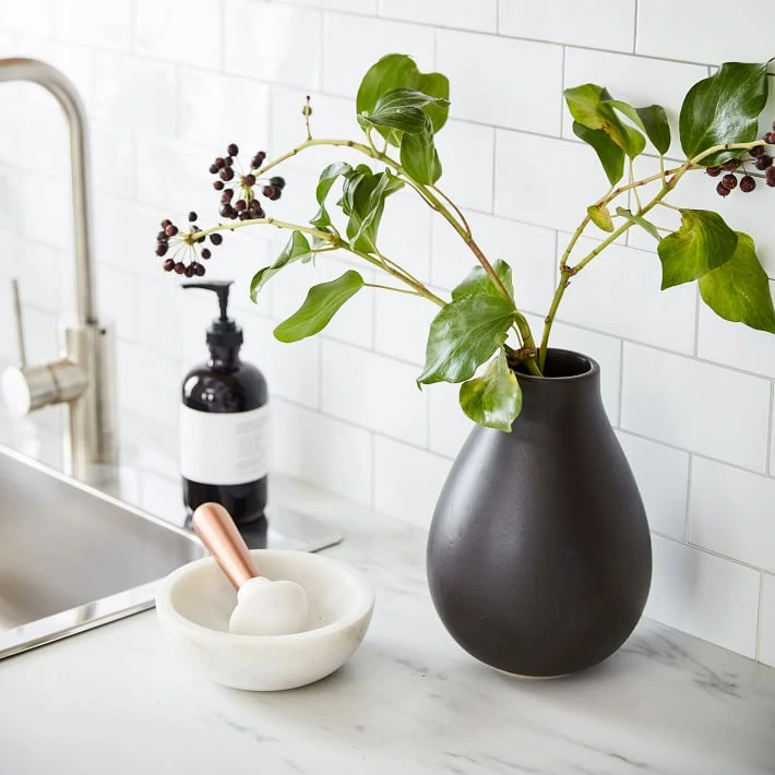 A matte black ceramic vase holding fresh greenery. Styled in a kitchen with marble countertops and white subway tile.