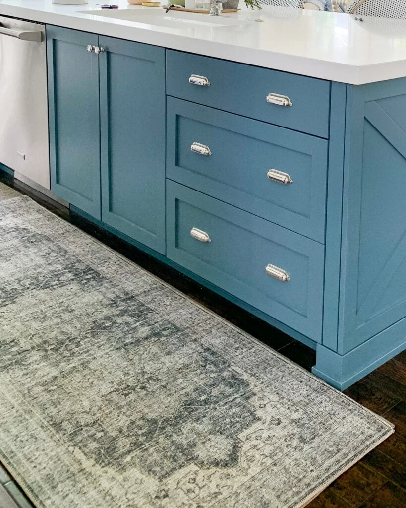 A blue kitchen island with silver bin pulls and a vintage style runner rug.