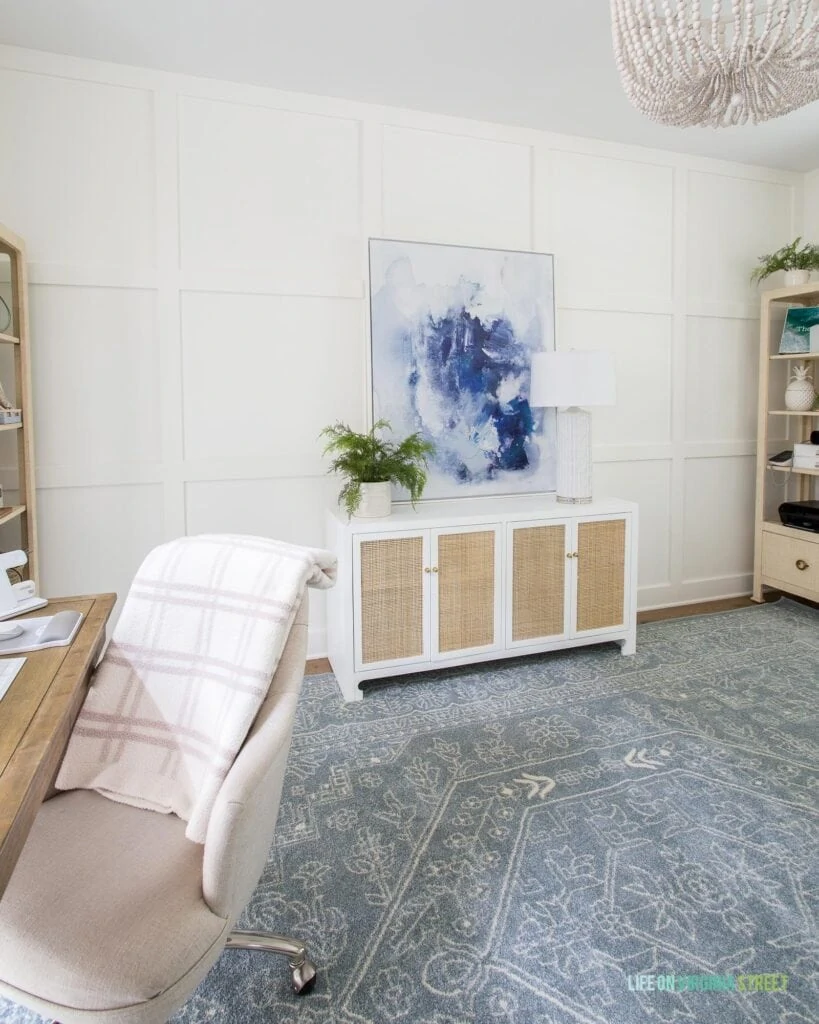 Home office with white board and batten grid wall, slate blue patterned rug, white and cane credenza, blue abstract art and linen desk chairs.