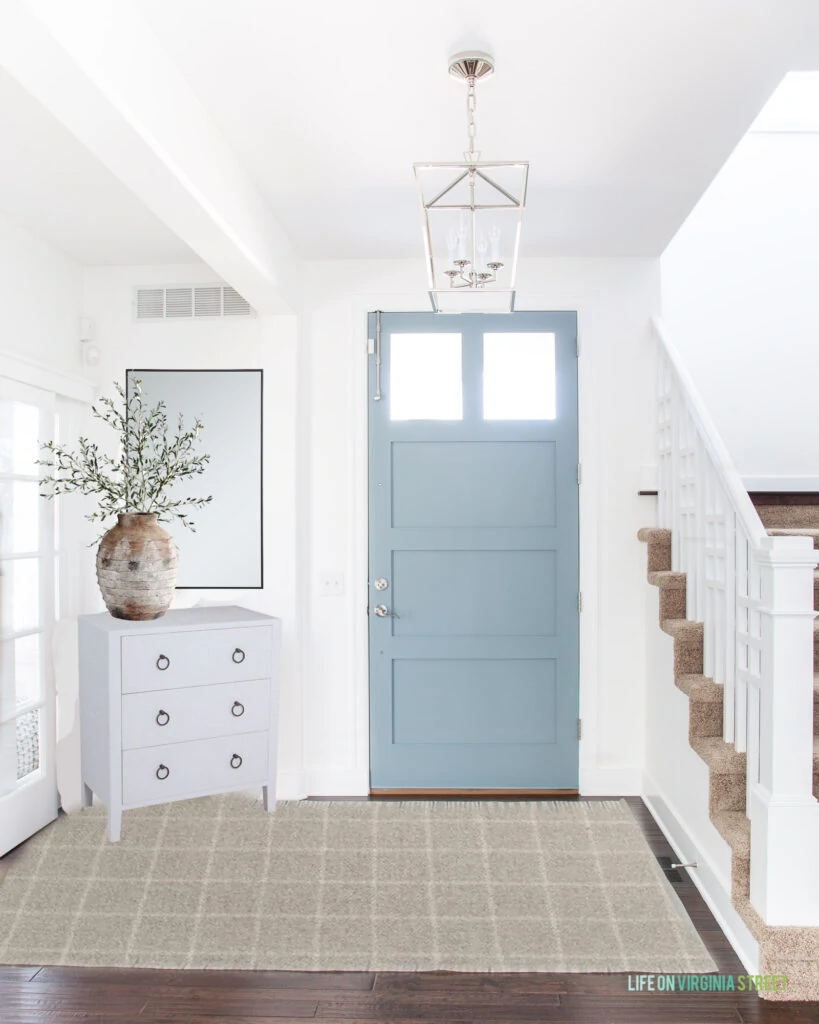 An entryway design plan with white walls, blue front door, silver chandelier, white raffia wrapped cabinet, a tall black mirror and a plaid rug.