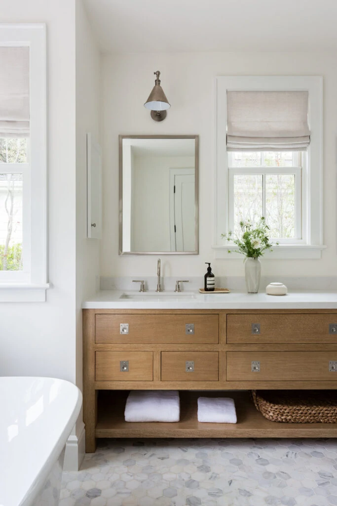 A beautiful bathroom with a long wood vanity, marble hex tile floors, a large tub, and windows with linen roman shades.
