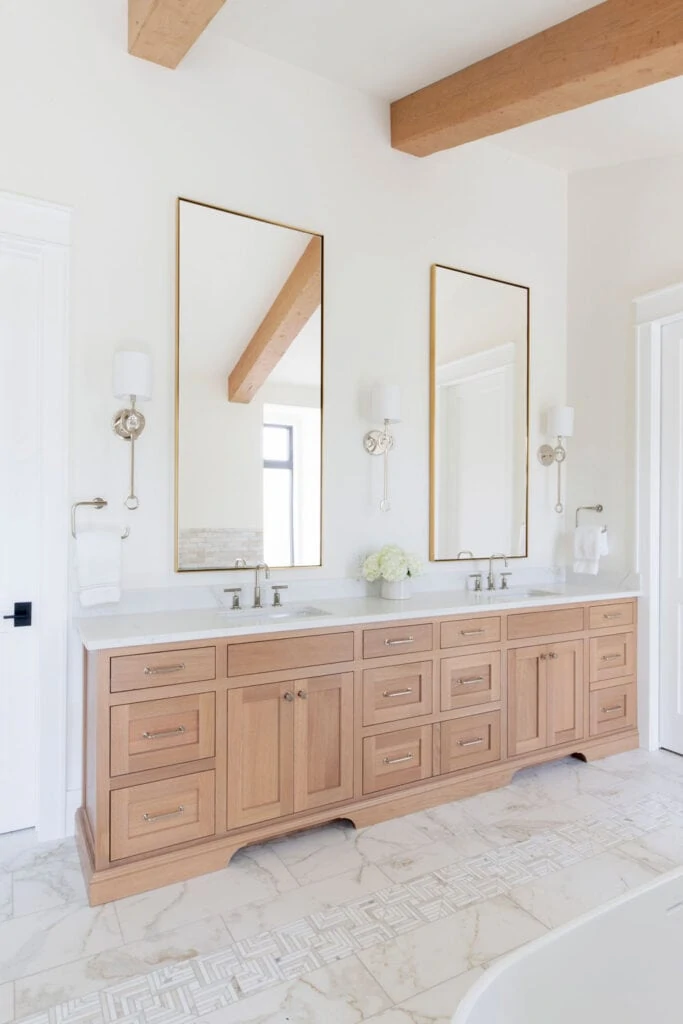 A large wood vanity in a bathroom with white walls, marble floors, wood beams, and tall brass mirrors.