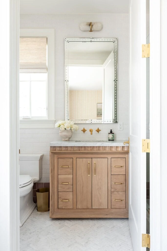 A cute girls' small bathroom ideas with herringbone marble floor, a fluted wood vanity, tall mirror, and a window over the toilet with a roman shade.