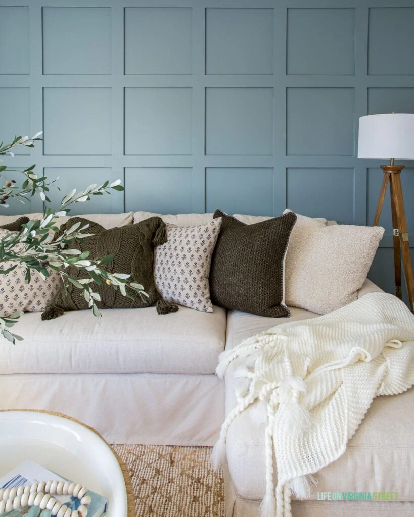 A moody board and batten grid wall (painted Benjamin Moore Van Courtland Blue) in a den with linen sectional, wood tripod lamp, and cozy pillows.