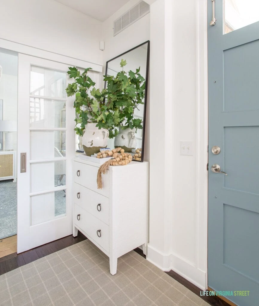 A white cabinet with drawers in an entryway with a blue front door, plaid rug, Tuscan vase filled with maples leaves, and a tall black leaning mirror.