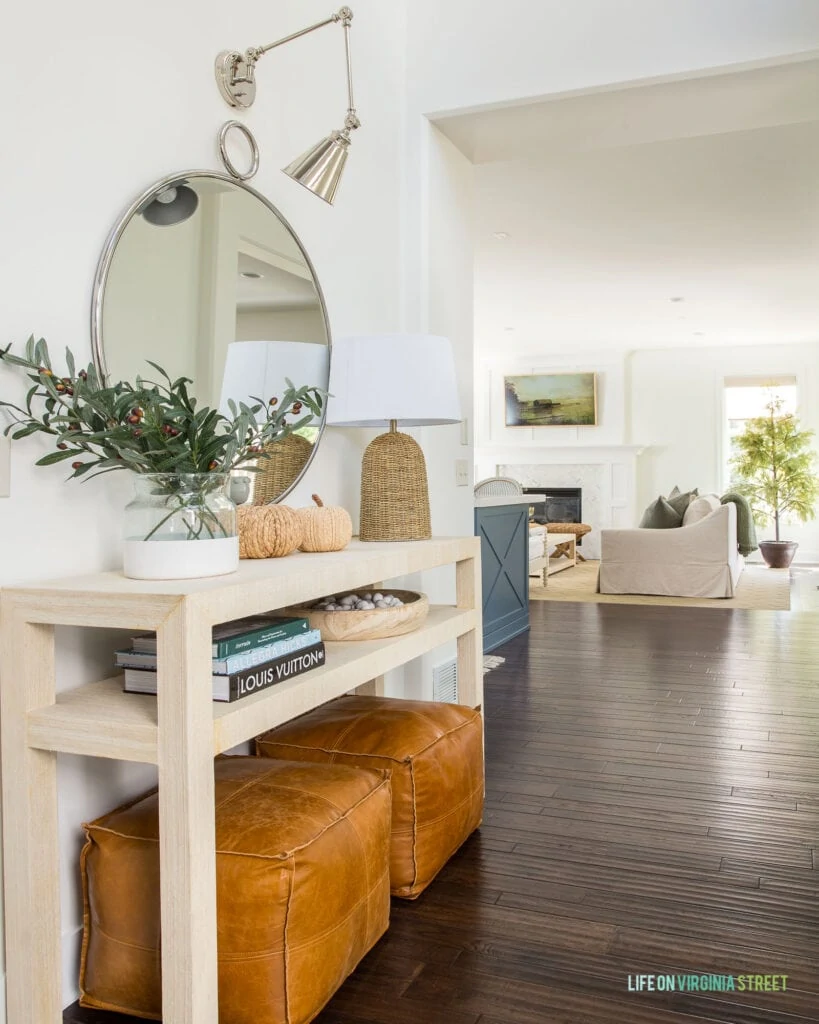 A raffia console table decorated for fall with leather poufs, faux olive stems, a round mirror, a silver swing arm sconce, raffia pumpkins, and a seagrass lamp.