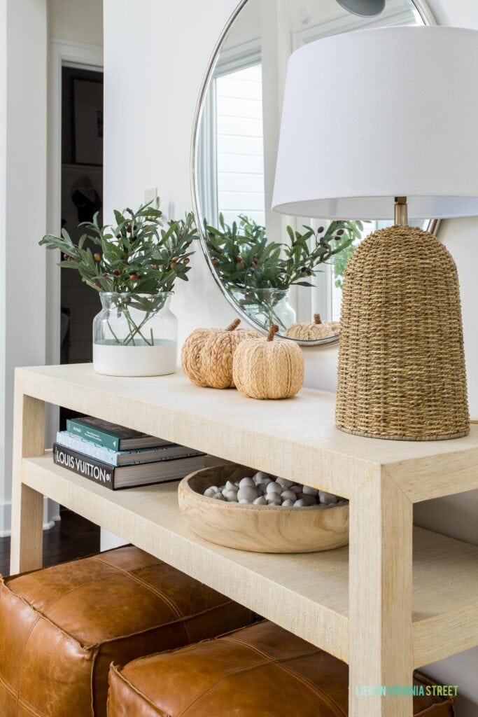 A raffia console table decorated for fall with leather poufs, faux olive stems, a round mirror, a silver swing arm sconce, raffia pumpkins, a wood bowl filled with white river rocks, and a seagrass lamp.
