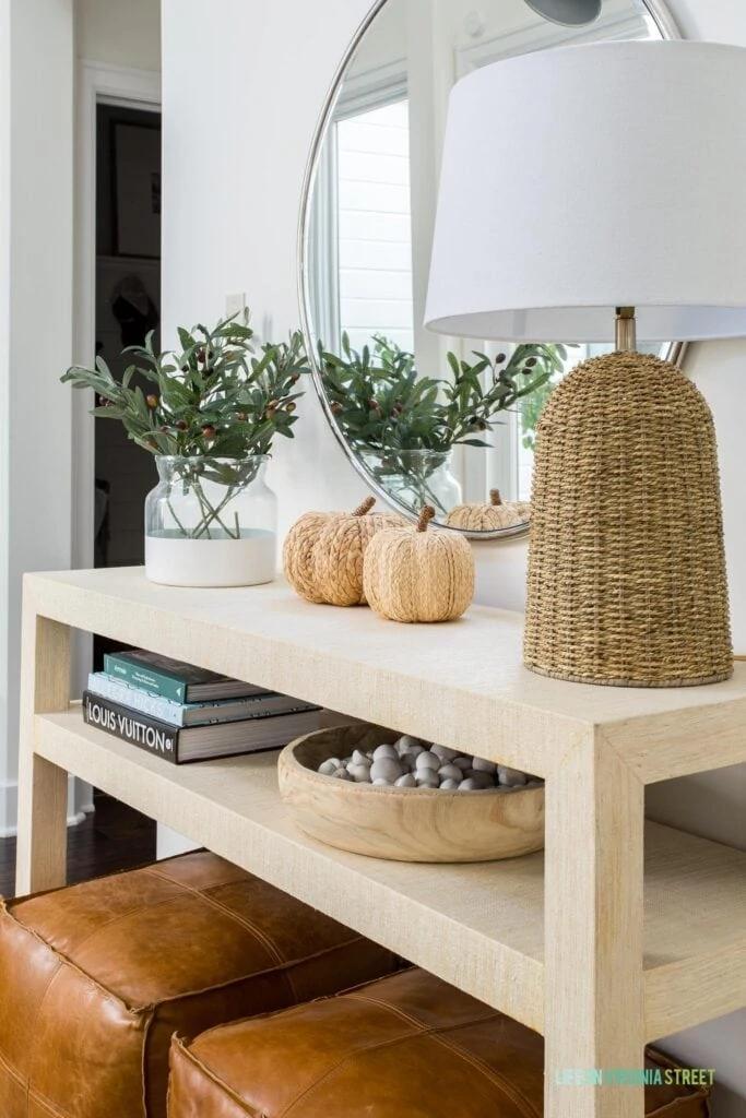 A raffia console table decorated for fall with leather poufs, faux olive stems, a round mirror, a silver swing arm sconce, raffia pumpkins, a wood bowl filled with white river rocks, and a seagrass lamp.