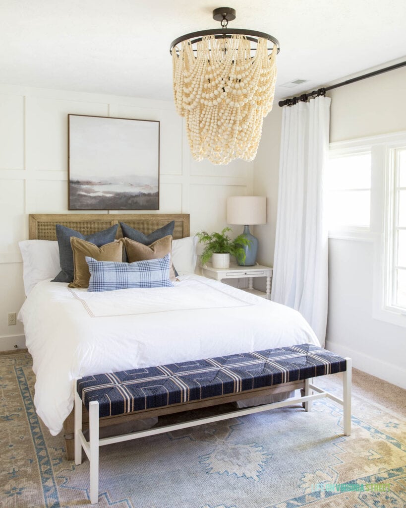 A guest bedroom with a bead chandelier, navy blue woven bench, white bedding, cane bed, abstract art, and large windows with white drapes.