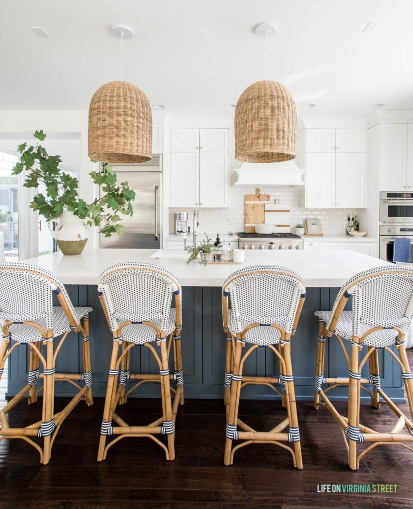 A beautiful fall kitchen with bistro style counter stools, woven pendant lights, dark blue island, and white kitchen cabinets with white beveled subway tile backsplash.