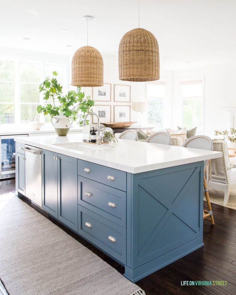 A beautiful kitchen with blue painted kitchen island, a neutral rug runner, basket pendant lights, large maple leaves, and a large window overlooking green landscaping.