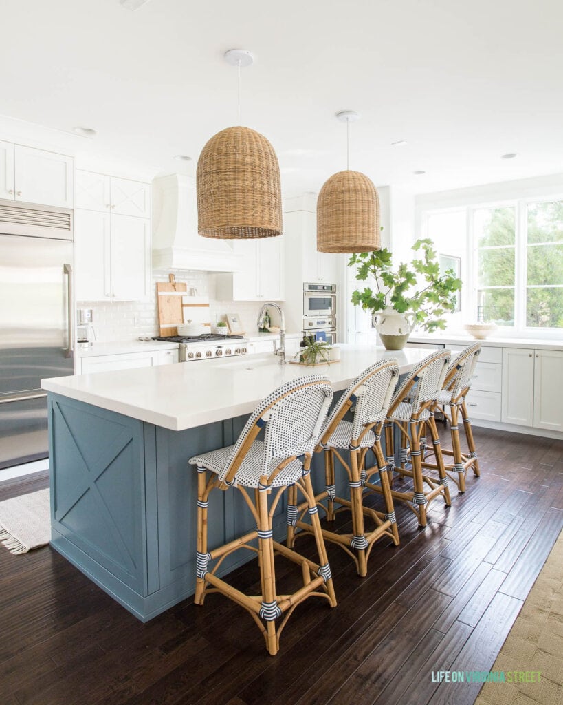 A fall coastal style kitchen with white cabinets, blue kitchen island, blue and white bar stools, woven pendant light fixtures, dark hardwood floors and a large built-in refrigerator.