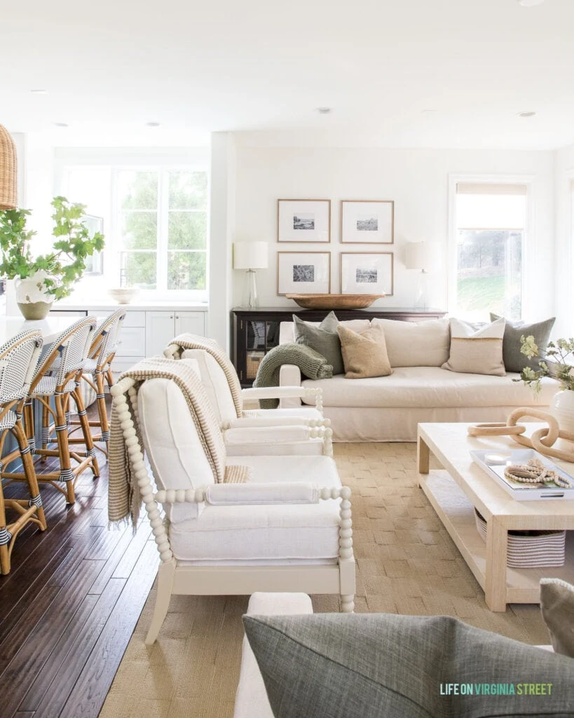 A fall living room with a photo gallery wall, linen sofa, white spindle chairs, sage green pillows, raffia coffee table and black console table.