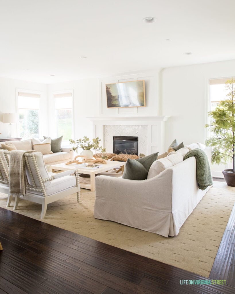A fall living room with Benjamin Moore Simply White walls, linen sofas, white spindle chairs, a raffia coffee table, a faux cypress tree and a Frame TV.