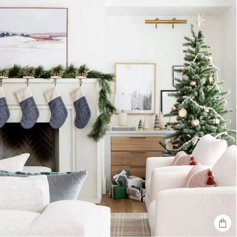 A beautiful white living room with garland on the white limestone mantel, with abstract art, blue colorblock stockings, a plaid rug and Christmas tree with metallic ornaments.