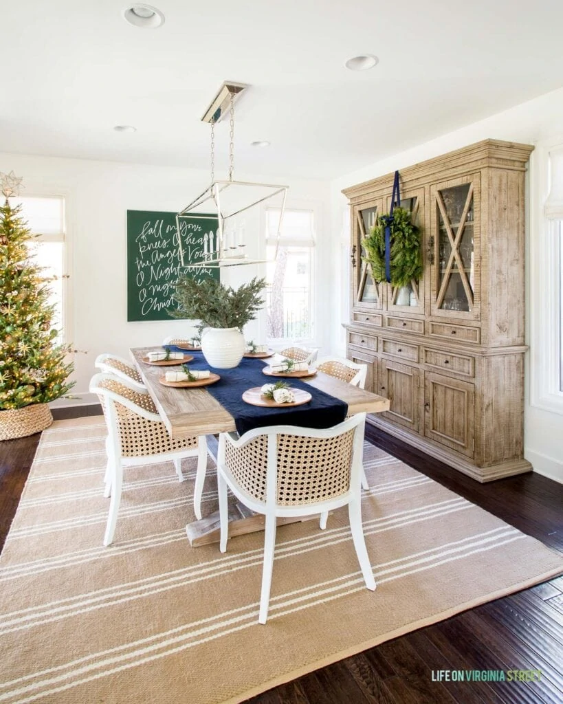 A neutral dining room decorated for Christmas with large carol artwork, a wreath, faux Christmas tree, navy blue table runner, and cane dining chairs.