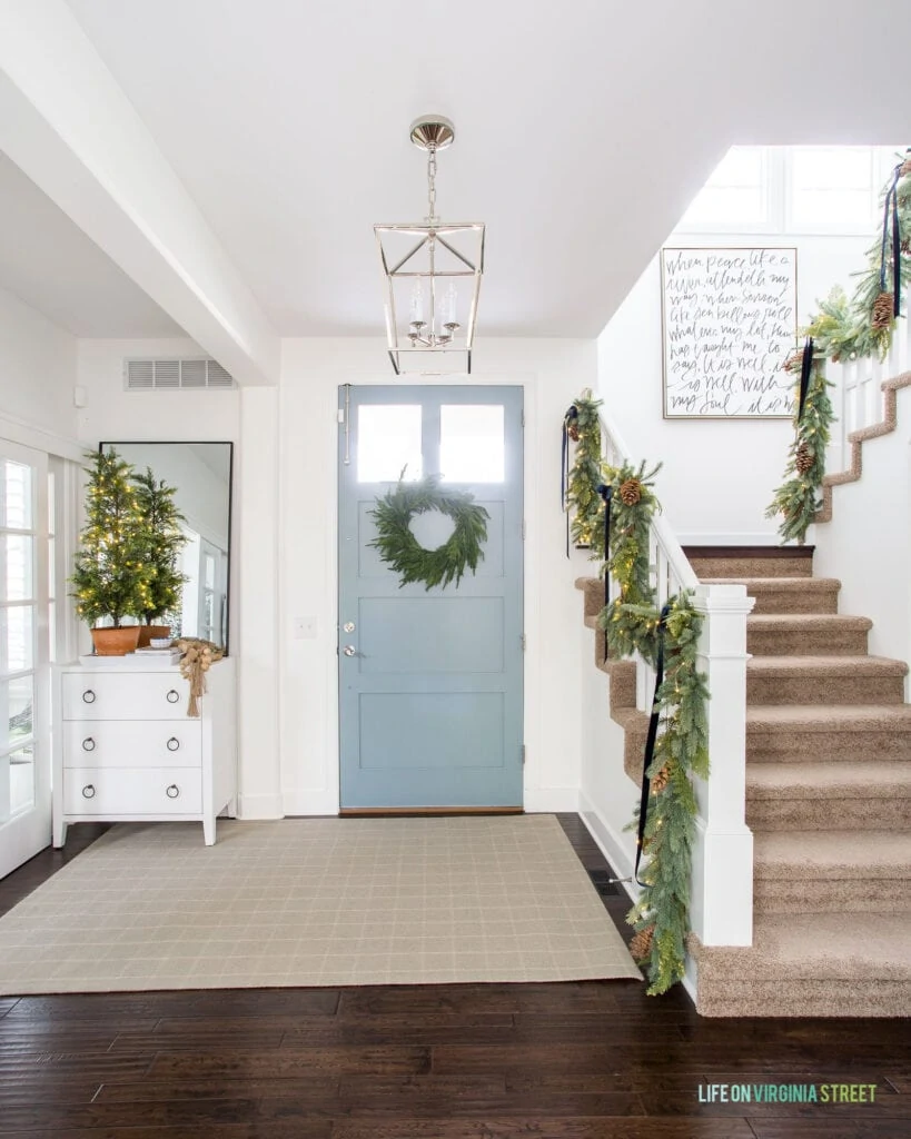 An entryway with white walls, blue gray front door, and plaid rug, decorated for Christmas with garland on the staircase and a wreath on the door.