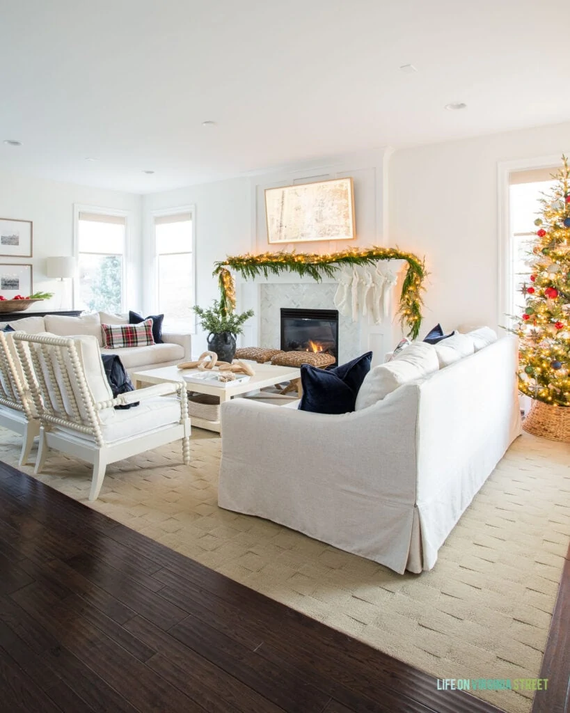 A white living room decorated for Christmas with a Christmas tree, navy blue and red ornaments, garland on the mantel with ivory stockings, and Stewart plaid pillows.