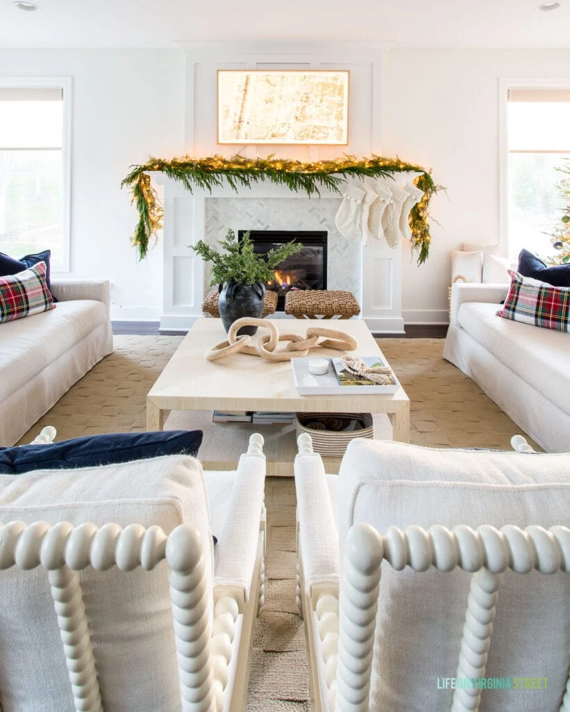 A Frame TV hanging over a Christmas mantel with ivory stockings and garland. You can also see a raffia coffee table, white spindle chairs and linen sofas with navy blue velvet and plaid pillows.
