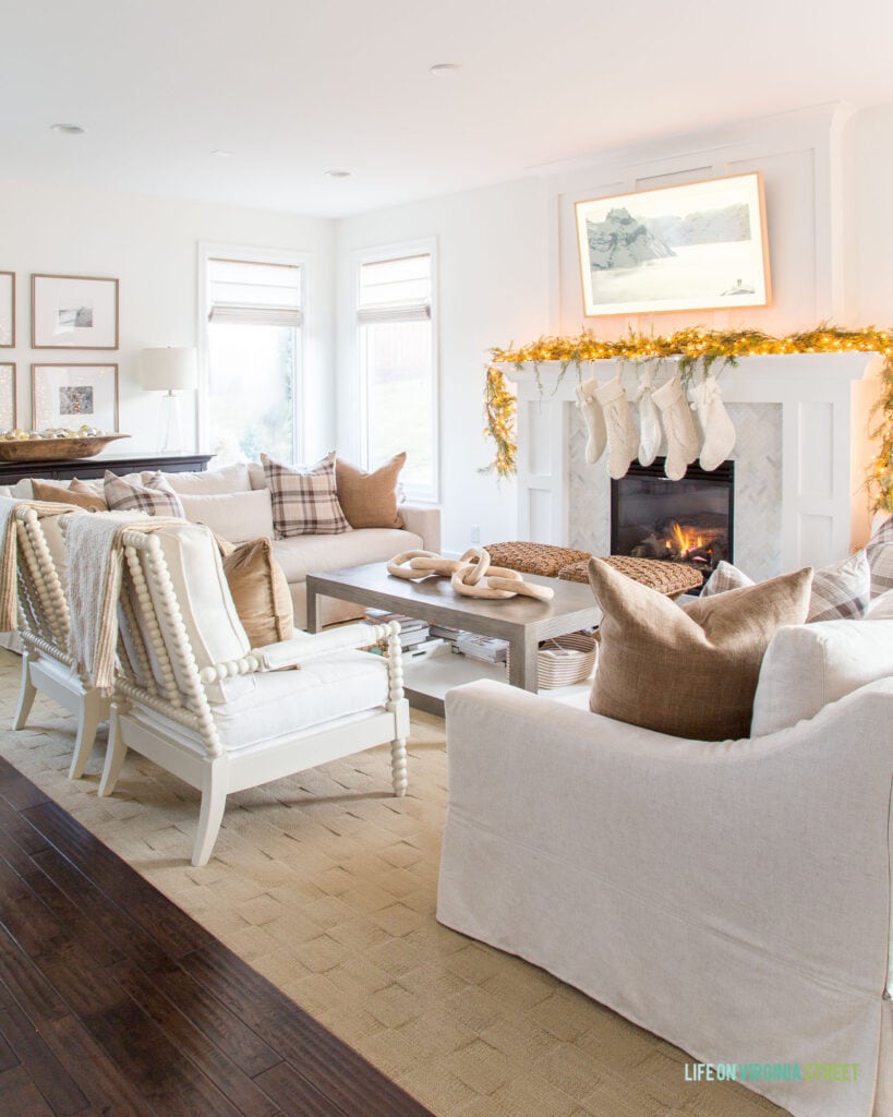 A neutral living room with Christmas decor including ivory knit stockings, brown plaid pillows, garland with lights and white spindle chairs.