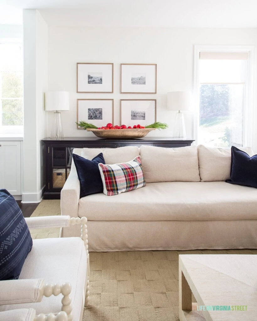 A linen sofa with navy blue velvet pillow, plaid lumbar pillow, a black console cabinet, wood gallery wall frames, and a wood dough bowl filled with bright red ornaments and faux evergreens.