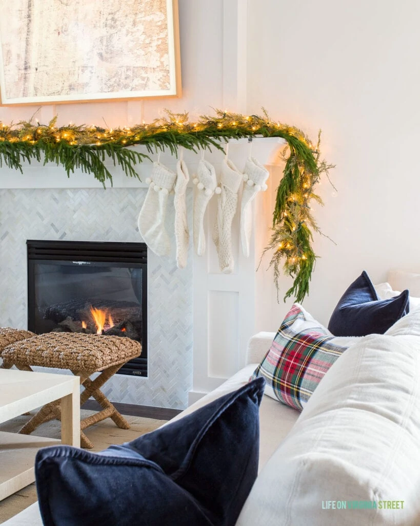 A Christmas mantel fireplace with green garland, twinkle lights, ivory woven stockings, rattan benches, and a linen sofa filled with navy blue velvet and plaid pillows.