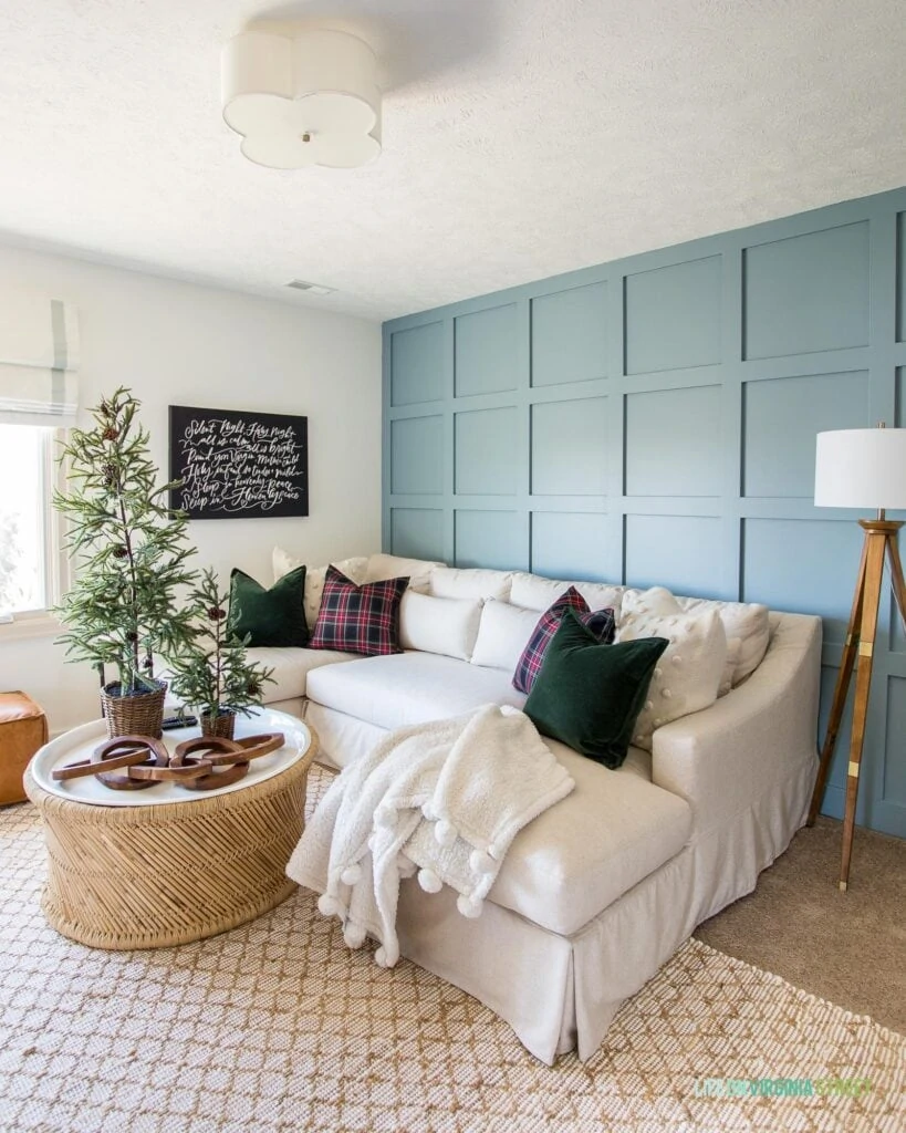 A den with a board and batten wall painted Benjamin Moore Van Courtland Blue. There is a linen sectional, bamboo coffee table, wood tripod lamp and Christmas decorations like mini Christmas trees, and plaid pillows.