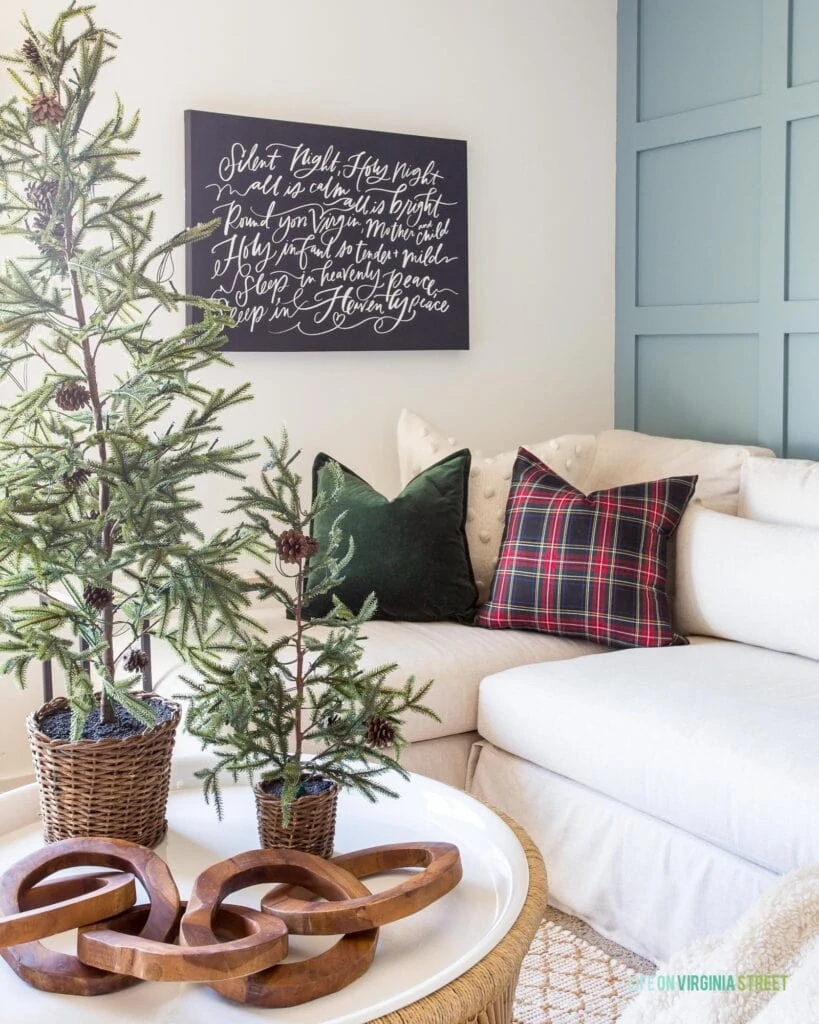 A cozy living room corner with a linen sectional, plaid pillows, a round coffee table, mini faux Christmas trees, wood link object decor, and a large 'Silent Night' canvas art piece.