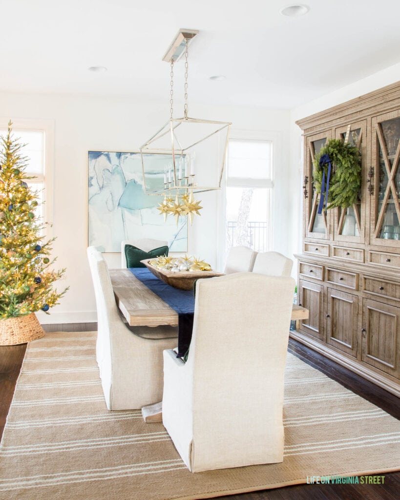 A Christmas dining room with camel striped rug, linen dining chairs, tall wood hutch, a natural style Christmas tree and gold stars hanging from the chandelier.