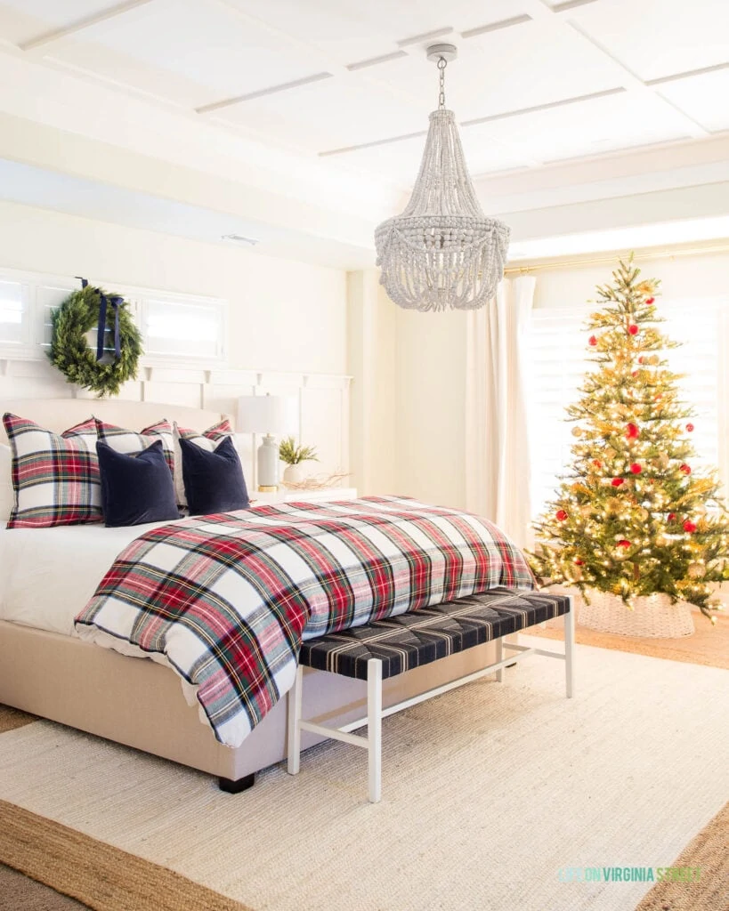 A bedroom decorated for Christmas with red plaid bedding, a natural style Christmas tree, white rope tree collar, jute rug, wood bead chandelier, and a wreath with navy blue velvet ribbon.
