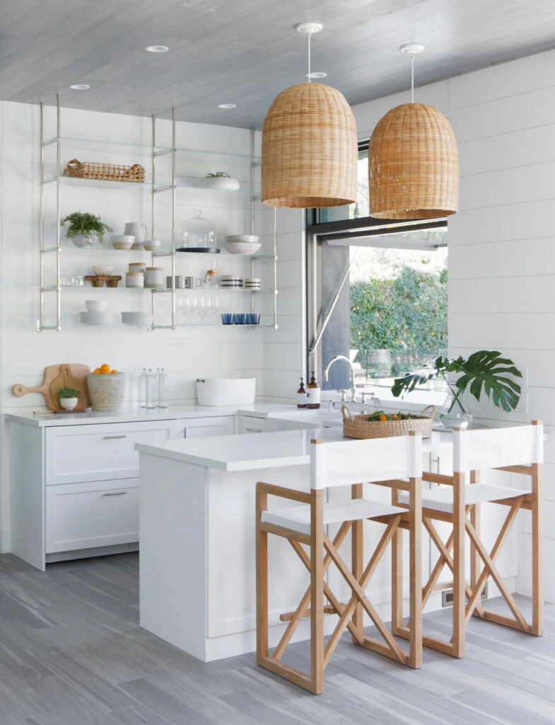 A pool house kitchen with small kitchen area, basket pendant lights, metal and glass hanging shelves, and a serving window over the sink that leads outside.