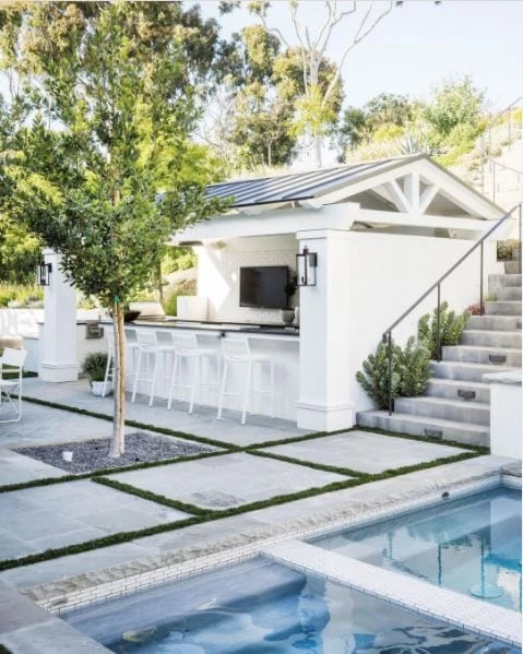 A gorgeous pool house idea with a white structure, metal roof, black countertop, TV, and bar stools. Love the pool and checkered landscaping as well.