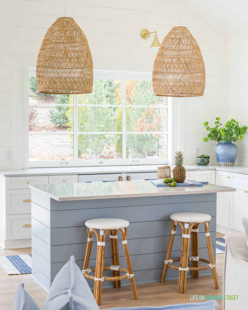 White woven backless counter stools with rattan legs in a kitchen with a large window, basket pendant lights, a blue shiplap island and white shiplap walls.