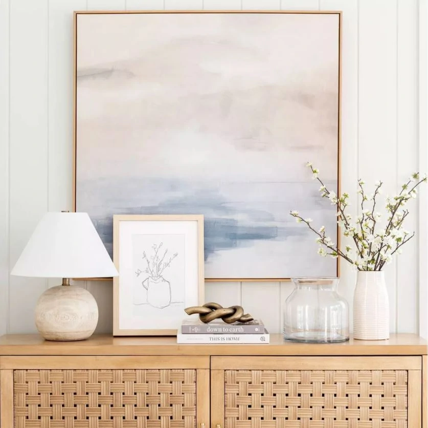 A gorgeous entryway console table vignette with pastel abstract art, a round wood lamp, metal knot decor, dogwood flowers in a vase, and a woven console table.