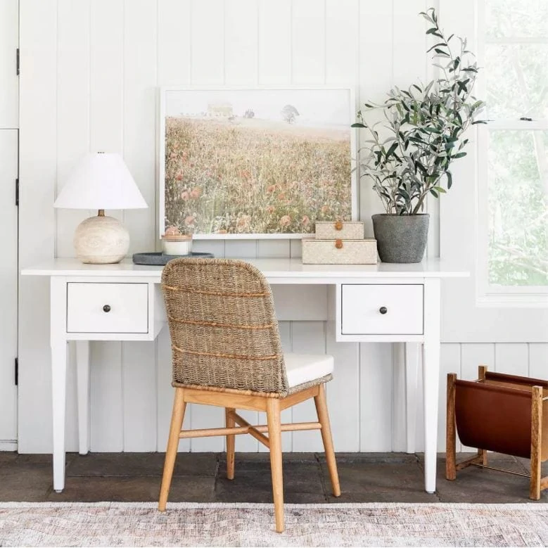 A cute white desk, woven chair, round wood lamp, wildflower art, and small olive tree in a home office space.